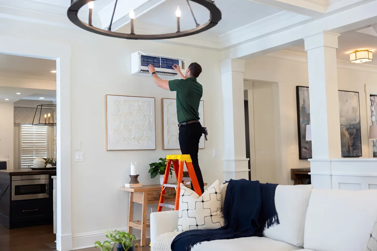 A Community Mechanical technician on a ladder services a wall-mounted ductless mini-split in a living room