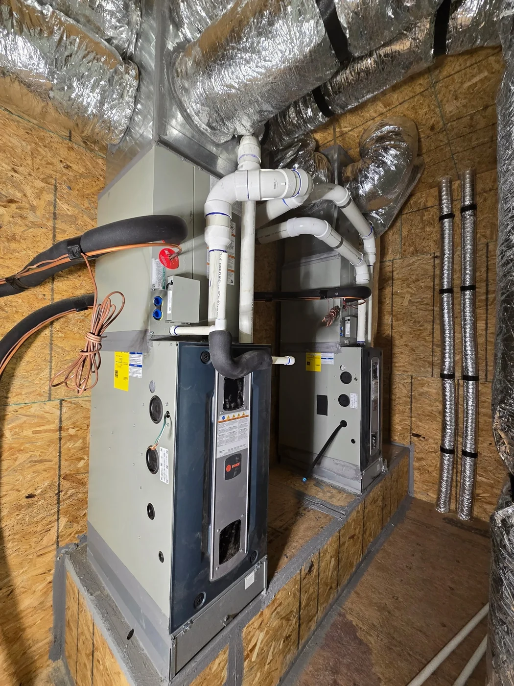 Two Trane gas furnaces side by side on a raised platform in a mechanical room, with PVC venting, condensate lines, and insulated flex ducts